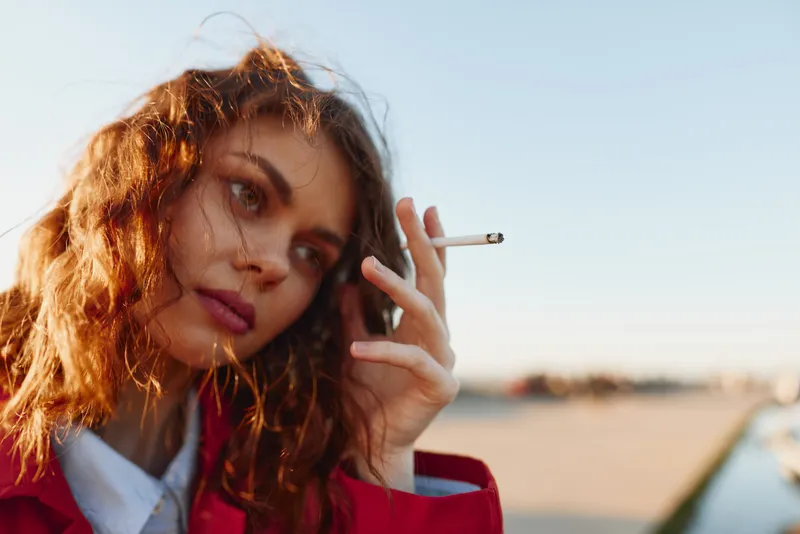 A young woman with red hair smokes a cigarette outdoors, wearing a red coat, captured in a candid, moody moment with soft sunlight.