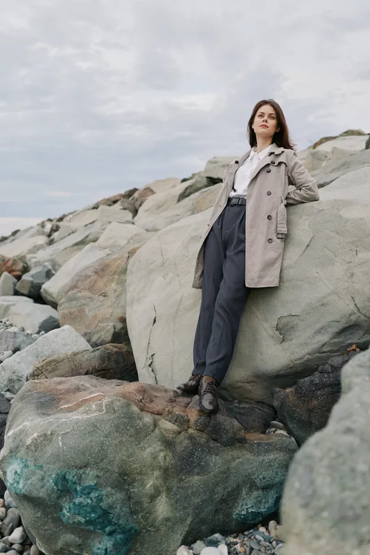 Young woman in casual trench coat and trousers stands confidently on rocky shore under cloudy sky. Concept of fashion, nature, confidence, and outdoor lifestyle with natural background.