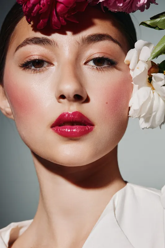Close-up of fashionable woman with glossy red lips, luminous skin, natural eyeshadow, and creative floral headpiece. Beauty, fashion, makeup, skin care conceptual studio portrait.
