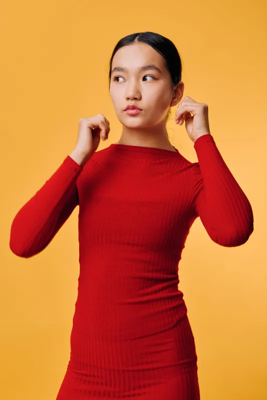Young woman wearing red dress posing and adjusting hair while looking to the side with thoughtful expression on orange background. Fashion style, portrait and beauty concept.