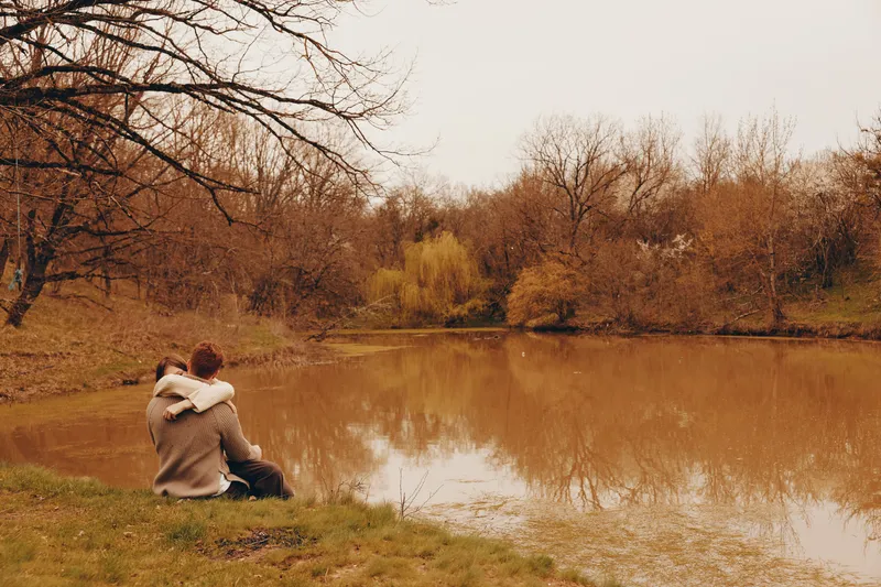 Couple sitting by a calm lake in autumn, embracing and enjoying the peaceful nature around them in warm sweaters on a cloudy day.