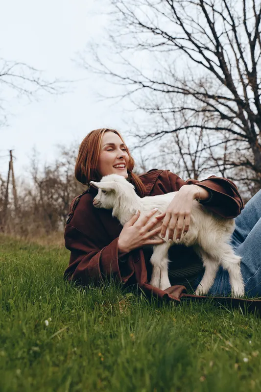 Young woman with red hair lies on green grass holding a white baby goat in her arms enjoying a peaceful moment outdoors in early spring with bare trees in the background.