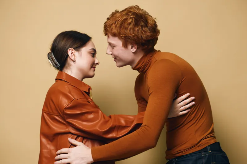 Couple embracing and looking at each other with smiles, wearing casual autumn clothes on beige background. Young man and woman in warm outfits sharing close moment.