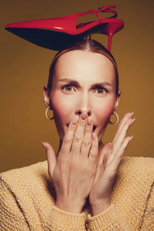 Woman with red high heel shoe on head covers mouth with hand, wearing yellow sweater and hoop earrings. Concept for surprise, fashion, and creative portrait in studio lighting.