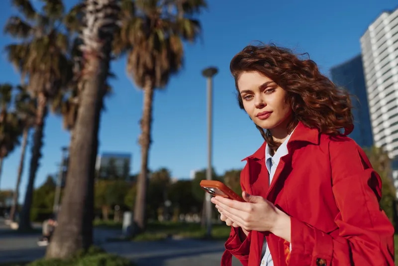 A young woman in a red trench coat stands outdoors on a sunny day, looking at her smartphone with a focused expression, palm trees and a city backdrop behind her.