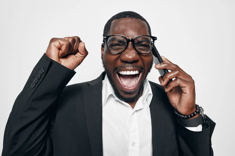 African American man with dark skin in glasses and suit talking on phone with excitement and fist raised for success and positive news in studio.