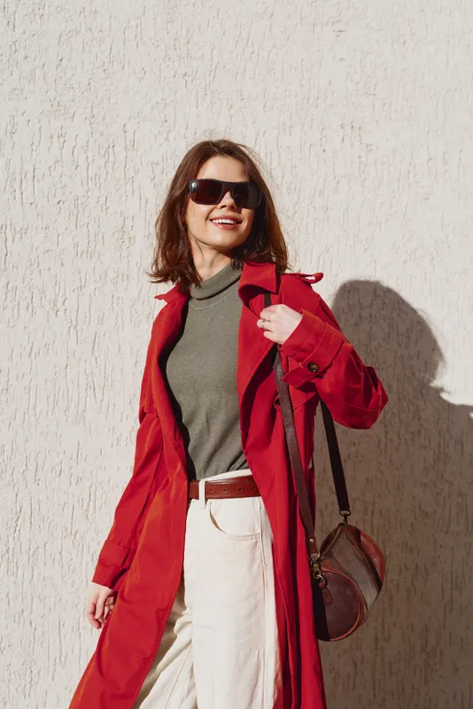 A stylish woman in a red coat and green top walks confidently along a light textured wall, wearing dark sunglasses and carrying a brown bag, exuding urban chic.