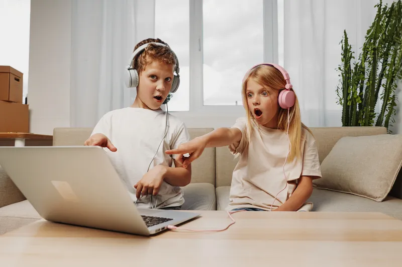 Two surprised children wearing headphones listen to music on a laptop at home, sitting on a couch. Concept of technology, entertainment, childhood, and digital leisure together.