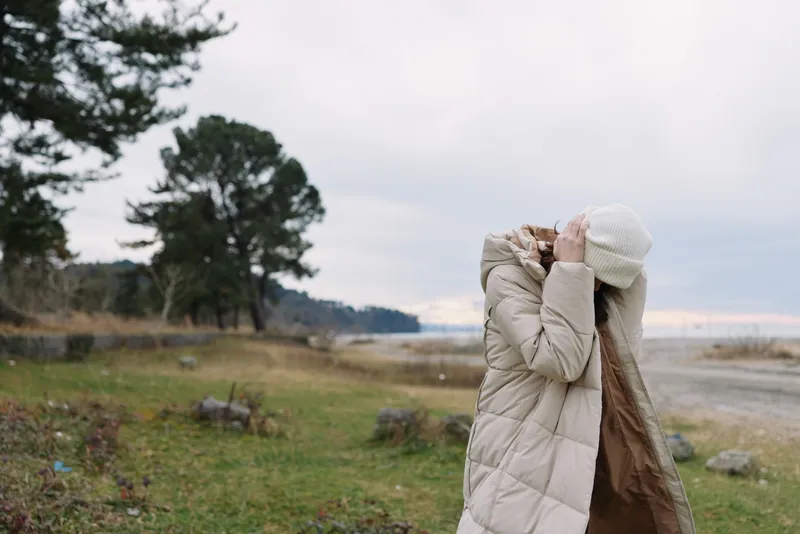 Outdoor woman in a beige winter coat and white hat adjusts her hood beside a foggy landscape with lake, trees and grass, conveying peaceful solitude and seasonal fashion in nature