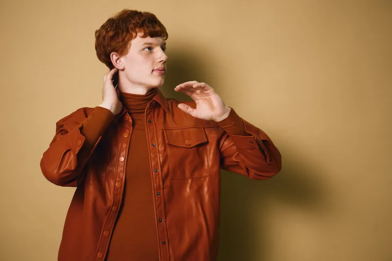 Young man with red hair wearing brown leather jacket and turtleneck standing against beige background looking sideways with hand near head, casual fashion portrait studio.