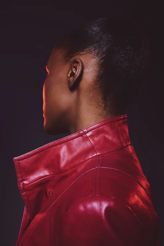 Portrait of a black woman with dark skin wearing a red leather jacket, shown from behind against a dark background. Concept of fashion, style, and modern elegance.