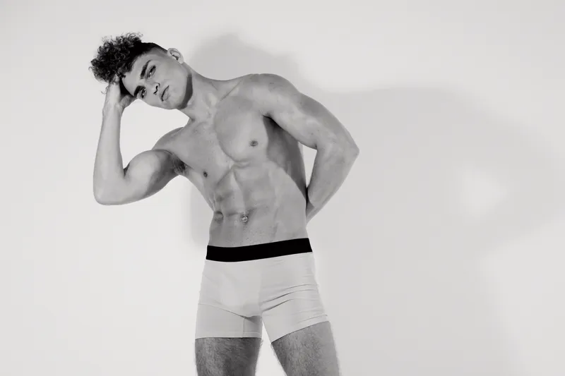 Young sporty male model posing shirtless in boxer briefs with one hand on head and serious expression on grey background in studio lighting.