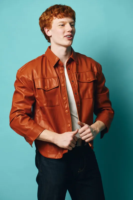 Young man with red hair wearing brown leather jacket buttoning shirt with casual style on blue background. Person standing, portrait with confident and relaxed facial expression.