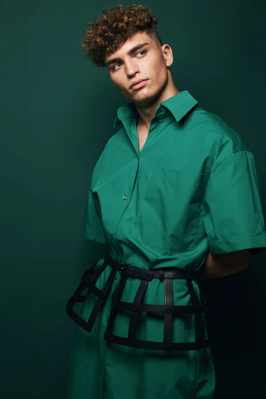 Young man with curly hair in green shirt and trousers standing against dark green background, fashionable male model posing with unusual black grid belt for editorial portrait and style.