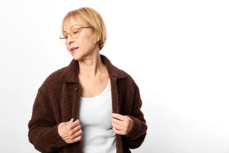 Woman portrait wearing brown jacket sweater, blond short hair, studio white background, adjusting warm casual clothing, soft light, calm thoughtful expression