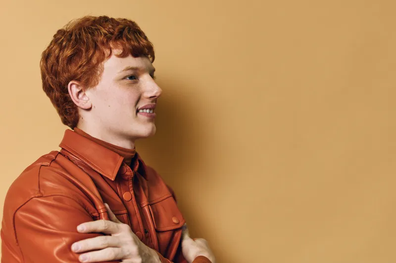 Young man with red hair and freckles smiling and looking sideways wearing brown leather jacket against plain beige background in studio portrait.