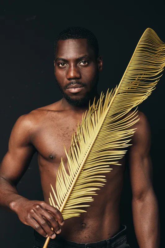 African man with dark skin and bare torso holding large golden palm leaf in creative conceptual visual style on black background. Artistic portrait with focus on texture and contrast.