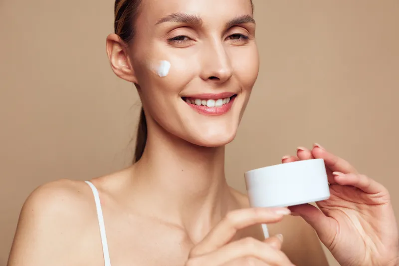 Smiling woman applying face cream with soft skin, natural makeup, holding white jar, skincare routine, healthy beauty, close up portrait, wellness concept