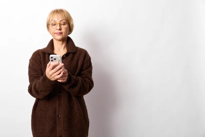 Elderly woman holding smartphone, technology communication, studio portrait with gray hair and warm sweater, calm smile while texting, white background and copy space