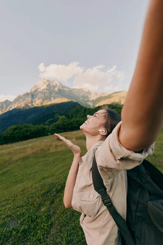 A joyful woman with a backpack enjoys nature with arms outstretched in a green field near mountains under a clear sky. Concept for lifestyle and freedom.