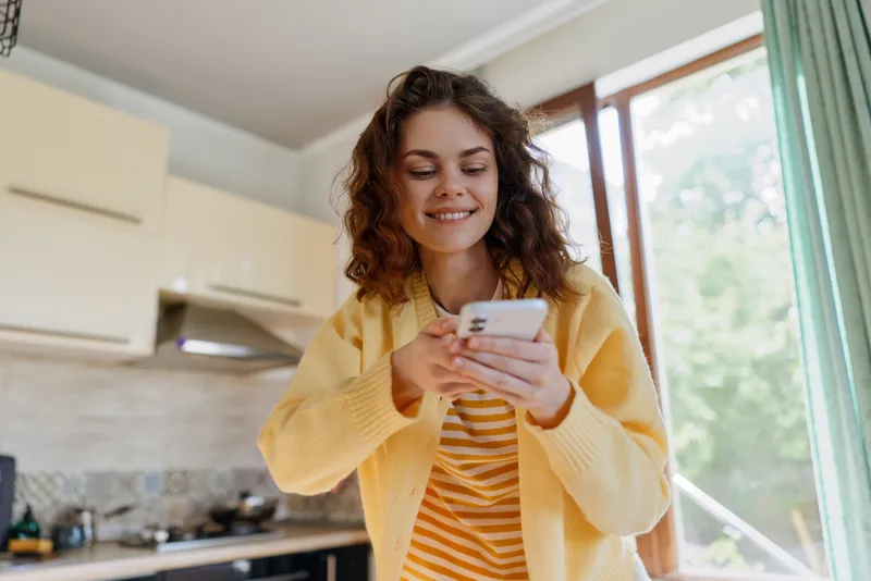 Smiling woman with curly hair using a smartphone in a bright kitchen. Casual home setting, warm daylight, relaxed mood, everyday technology, self expression, cozy atmosphere, morning routine.