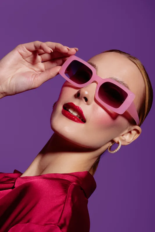 Stylish woman with pink sunglasses and red lipstick poses against purple background. Fashion portrait with glossy makeup and elegant earrings. Concept of glamour and modern style.