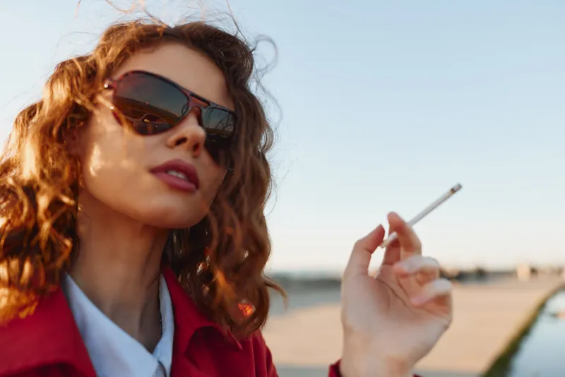 Young woman outdoors holding cigarette with blurred cityscape background, wearing sunglasses and red coat, natural light, casual pose, clear sky