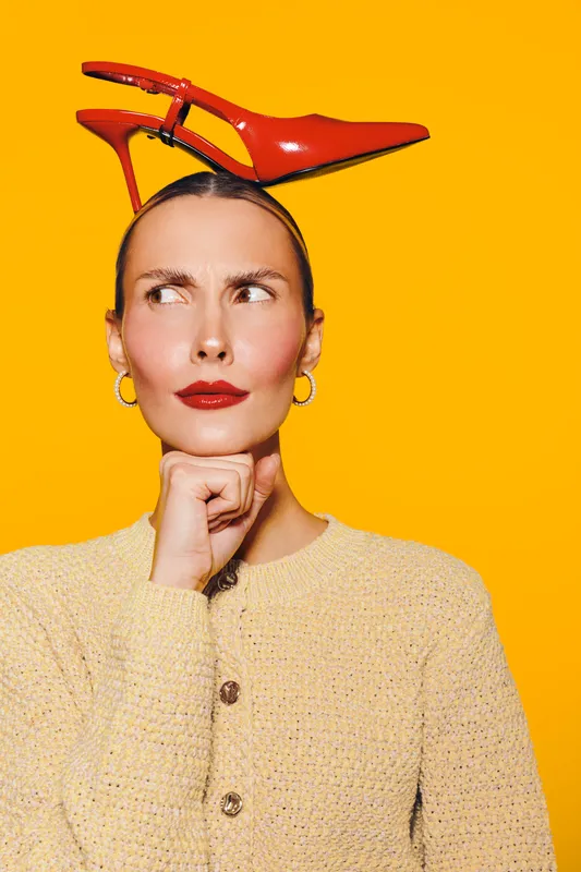 Young woman with red high heel shoe on head looks sideways with thoughtful expression. Bright yellow background and casual beige sweater create playful and quirky fashion concept.