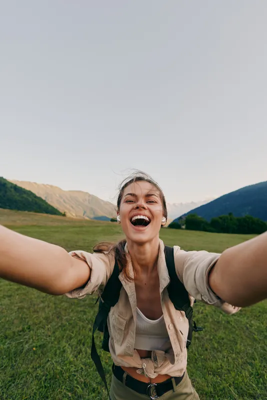 Cheerful woman outdoors, smiling with arms extended, sunlit meadow and distant mountains, casual adventure vibe, natural colors, carefree lifestyle moment