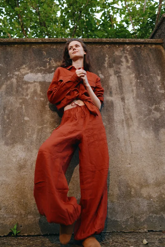 Woman outdoors fashion lifestyle jumpsuit red pose confident, standing against a weathered wall, with natural light and greenery, urban vibe, relaxed expression, bold color and texture