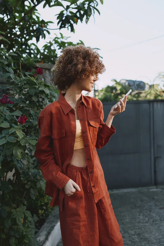 Woman in orange casual outfit stands outdoors, checking her smartphone, embracing a relaxed lifestyle, natural light, greenery in the background, candid moment of everyday fashion