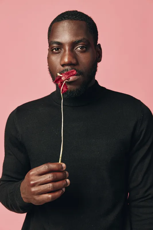 African man in black turtleneck holding red flower near lips with serious expression on pink background. Creative conceptual visual style portrait with dark skin and thoughtful mood.