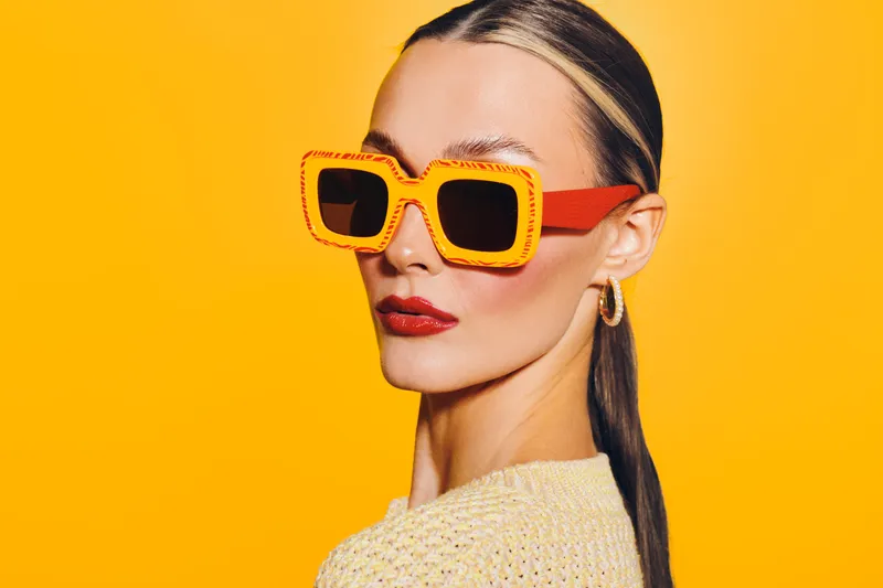 Stylish woman with slicked back hair wearing oversized orange sunglasses and red lipstick poses against bright yellow background. Concept of fashion, summer style, and confident attitude.
