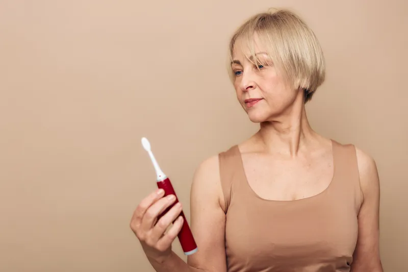 Mature woman with short blonde hair examines electric toothbrush in beige tank top against neutral background. Concept of dental care, personal hygiene, and healthy lifestyle for seniors.