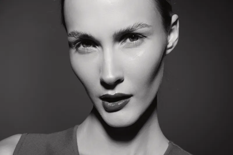 Close-up portrait of a young white woman with pale skin and sharp facial features looking confidently at the camera in black and white studio lighting.