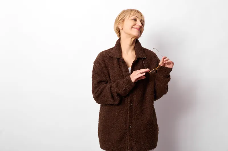 Woman studio winter coat smiling blonde portrait on white background, relaxed female posture, adjusting jacket collar, gentle laugh, soft studio light, neutral clean backdrop