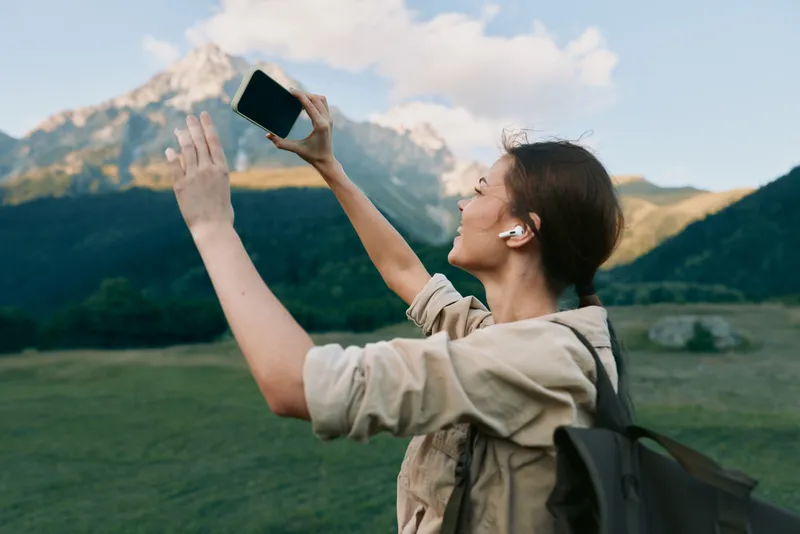 Woman with backpack outdoors in a mountainous landscape reaching toward a smartphone, wearing wireless earphones. Focus on lifestyle travel wellness and self expression bright airy natural light