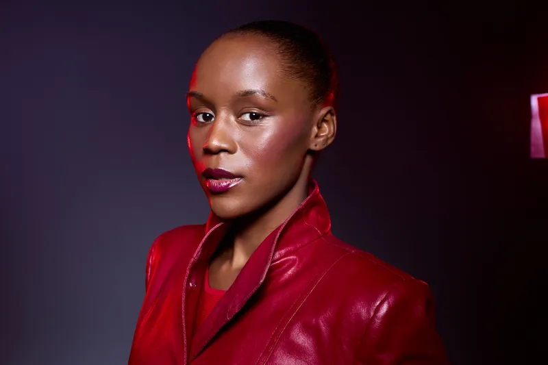 Portrait of a confident black woman with dark skin wearing a red leather jacket, looking at camera with soft lighting and dark background. Concept of fashion, beauty, and empowerment.