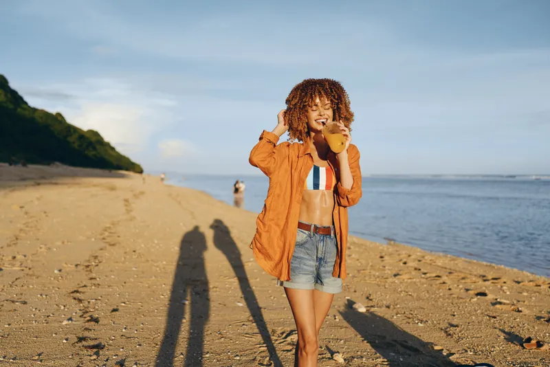 Young woman on sandy beach wearing orange jacket and denim shorts drinking juice with straw, sunny day, relaxed mood, ocean in background, casual summer outfit