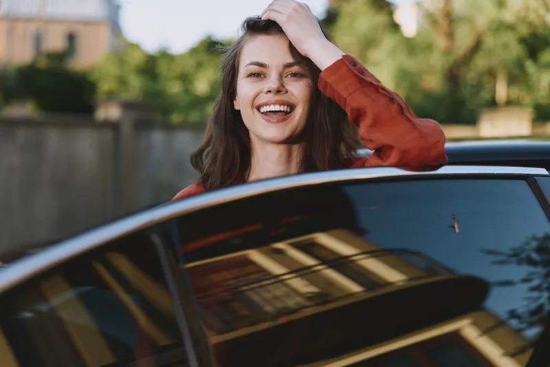 Cheerful woman outdoors, smiling broadly while leaning from a car window, sunlit moment, casual pose, natural colors, urban background, carefree lifestyle
