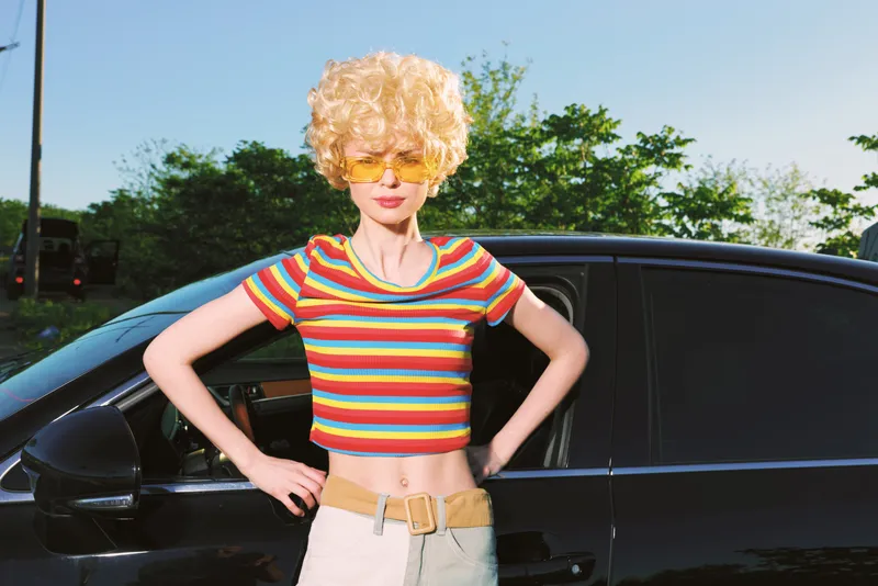 Young woman with curly blond hair wearing a rainbow crop top and sunglasses stands beside a dark car on a sunny day, radiating retro confidence.