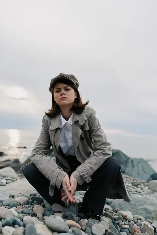 Young woman in vintage coat and cap poses confidently on rocky beach with ocean and cloudy sky in background. Concept of fashion, outdoor, autumn style, and natural beauty.