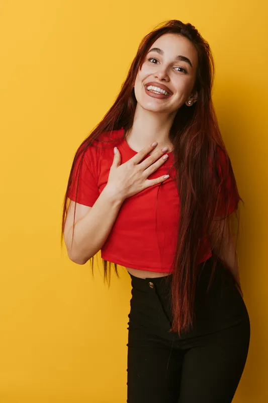 Young woman with long hair smiling and holding hand on chest against yellow background. Casual outfit with red shirt and black pants, happy expression and friendly gesture.