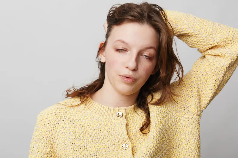 Young woman in yellow knitted sweater stretches with eyes closed and hand behind head. Studio portrait on grey background. Concept of relaxation, tiredness, and casual fashion.