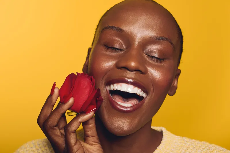 Happy smiling black woman with dark skin holding red rose near face on yellow background. Concept of joy, beauty, self-love, and natural skincare with positive mood in studio lighting.
