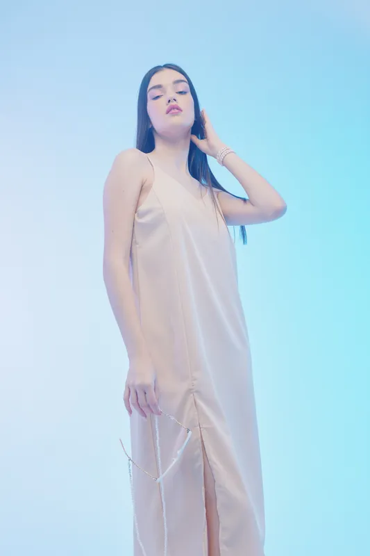 Young woman in a beige satin dress poses confidently against a soft blue background. Her long dark hair and minimal makeup highlight natural beauty and fashion elegance.