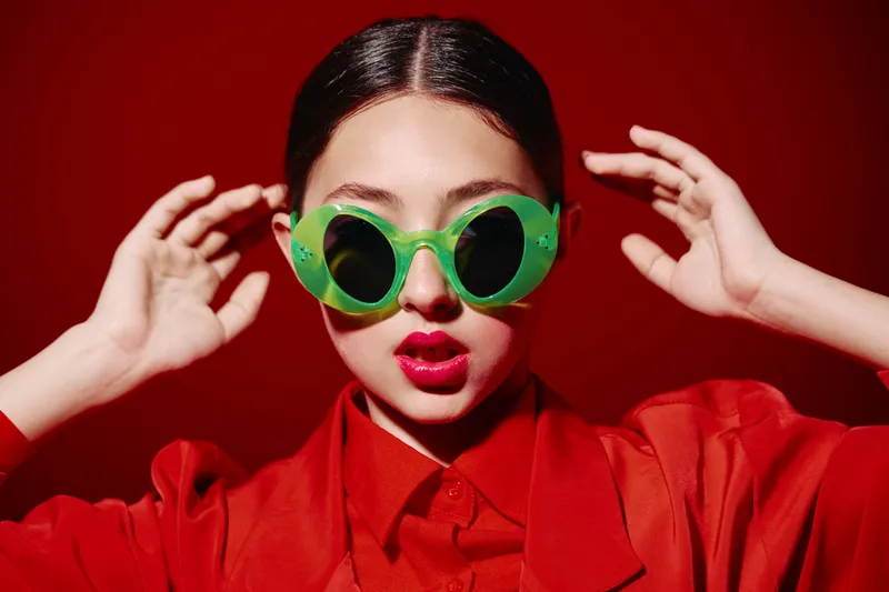 Young woman with bright makeup wearing large green fashion sunglasses and red outfit against red background. Creative concept for beauty, fashion, and editorial style shoot.