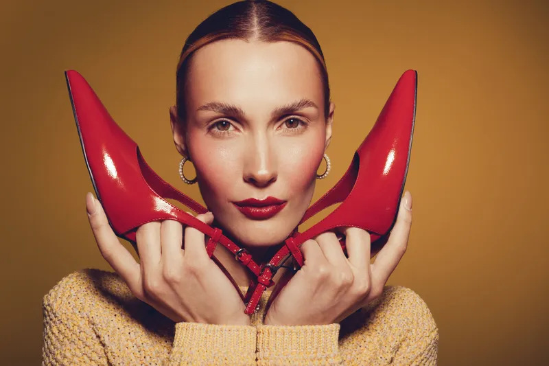 Portrait of a stylish woman holding red high heel shoes near her face on a warm brown background. Concept of fashion, beauty, elegance, and modern style with confident expression.
