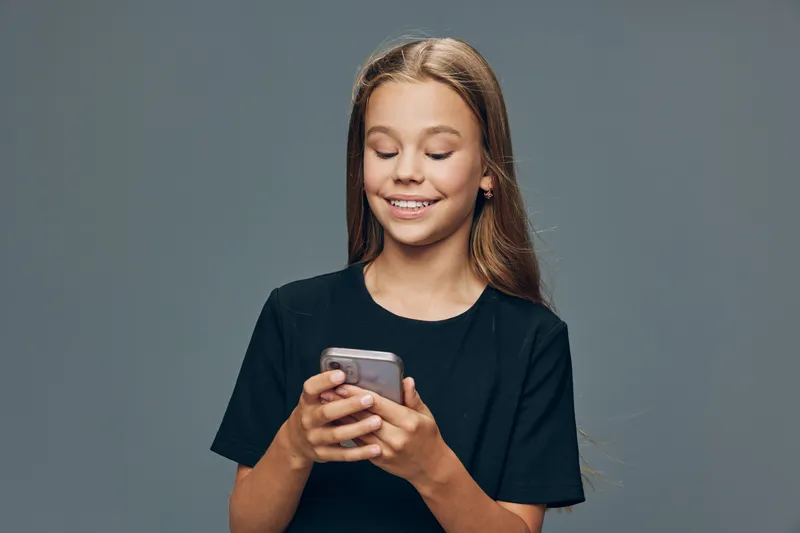Girl with long hair smiling and looking at smartphone in hands, casual black t shirt, isolated on gray background, enjoying mobile phone use and digital communication.