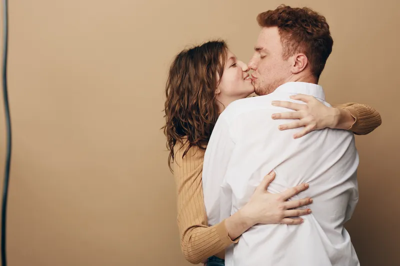 Young couple embraces and kisses tenderly against beige background. Woman in brown sweater and man in white shirt share intimate moment of love and affection. Romantic relationship concept.